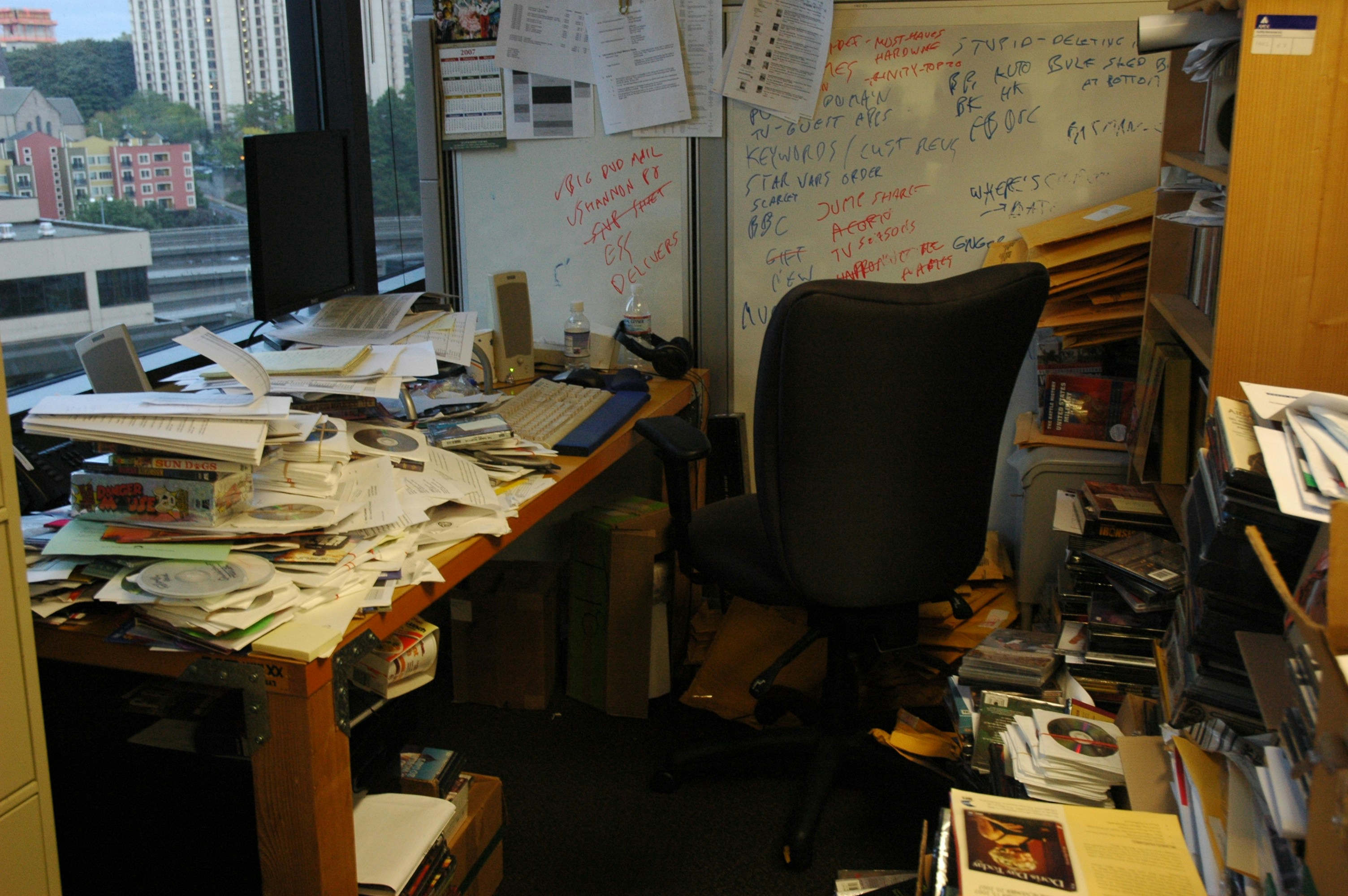 A cluttered office workspace filled with large piles of papers, books, CDs, and miscellaneous items on the desk and floor, with a computer monitor, keyboard, and an empty office chair in front of a whiteboard covered in handwritten notes.